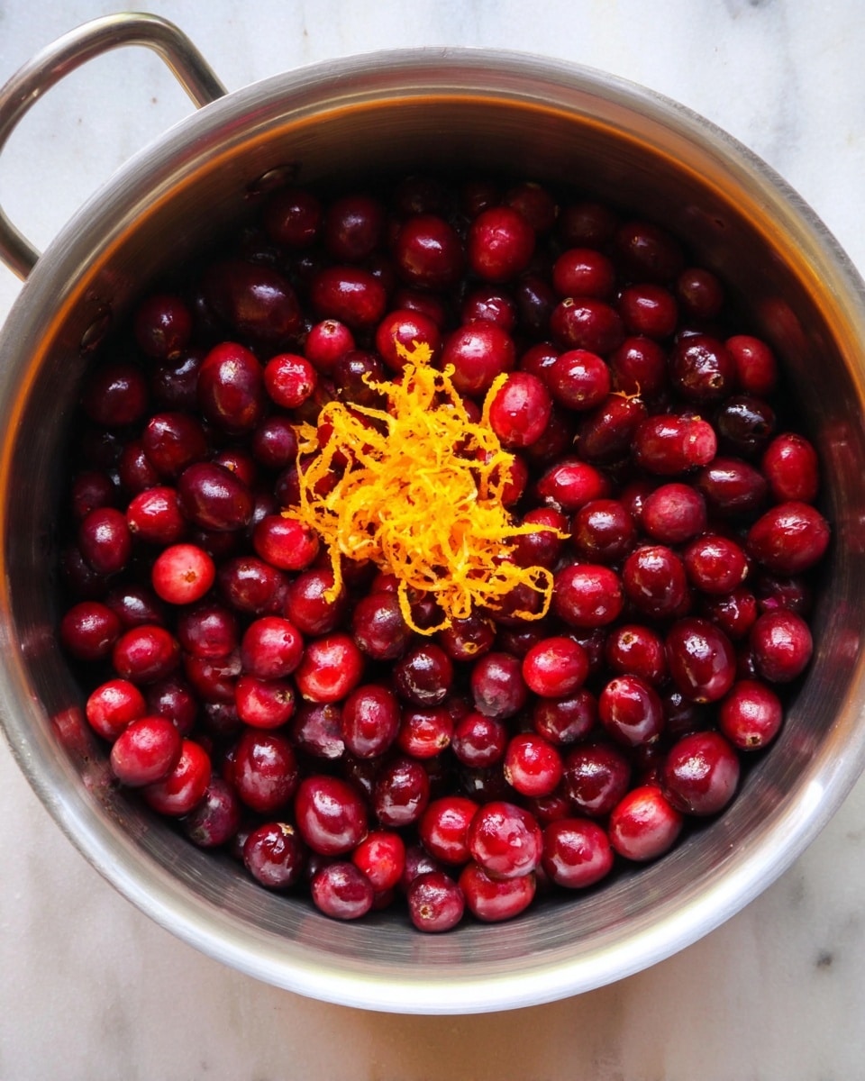 A shiny silver pot is filled with many small, round, deep red cranberries that cover the entire bottom and sides. On top of the cranberries, in the center, is a small pile of bright orange zest with a fine, curly texture. The pot sits on a white marbled surface. photo taken with an iphone --ar 4:5 --v 7