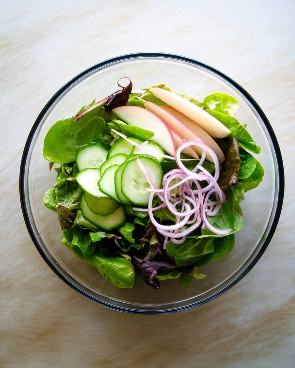 A clear glass bowl sits on a white marbled surface filled with a fresh salad. The bottom layer is made of mixed green leafy lettuce with different shades of green and some purple leaves. On top of this is a layer of thinly sliced light green cucumber slices arranged in small clusters. Scattered on the salad are thin, pale purple rings of red onion adding light color contrast. Additionally, there are thin slices of pale pink apple with a slight red edge layered on one side. The overall look is fresh, light, and colorful. Photo taken with an iphone --ar 4:5 --v 7
