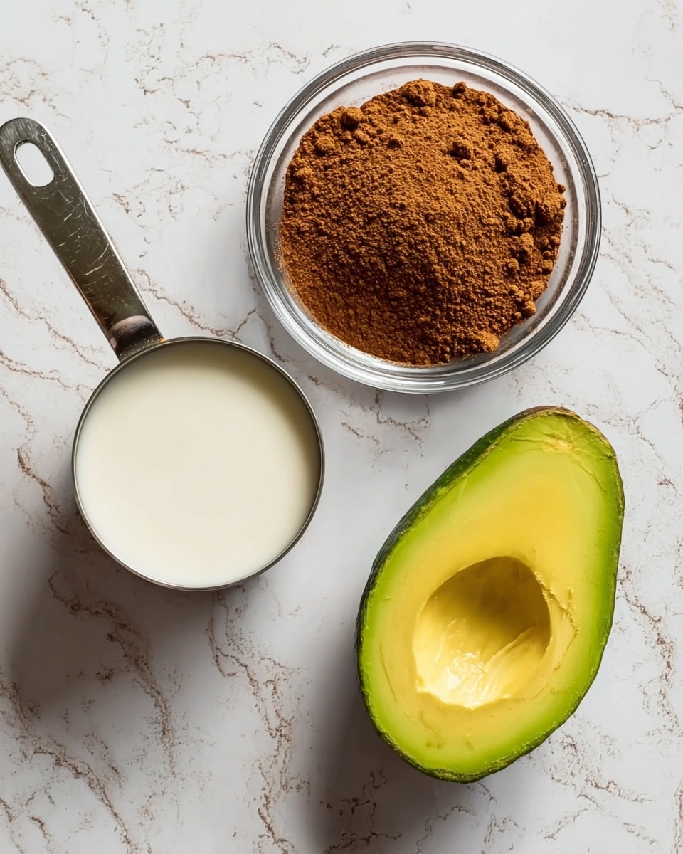 The image shows four main items placed on a white marbled surface. At the bottom right, there is a half of a ripe avocado with a smooth, bright green and yellow inside texture and a dark green skin around it. Just above the avocado is a clear glass bowl filled with a brown powdery spice that is loosely piled up with small clumps. To the top left is a small stainless steel measuring cup filled with a white liquid, likely coconut milk or cream, with a shiny handle extending outward. Part of a clear glass container is visible at the bottom left edge of the image. photo taken with an iphone --ar 4:5 --v 7
