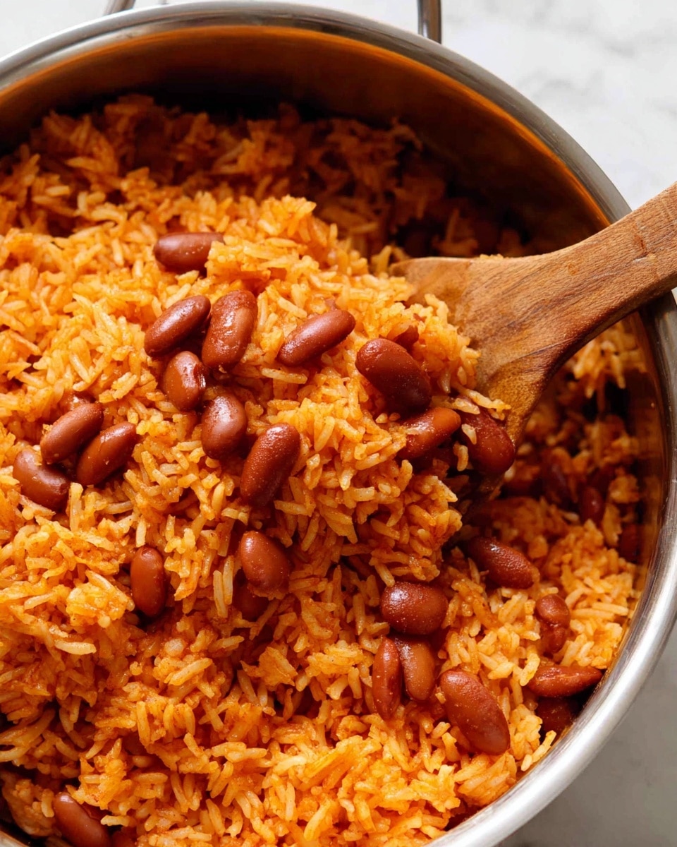 A white bowl filled with orange rice mixed with brown beans, topped with small green herb leaves scattered across. A fork rests inside the bowl on the left side, with part of a woman's hand holding it. In the background, slightly out of focus, is a white bowl full of fresh green leafy vegetables, a bottle of red hot sauce, and a glass of yellow liquid. The bowls and items are placed on a white marbled surface with a blue cloth napkin nearby. photo taken with an iphone --ar 4:5 --v 7