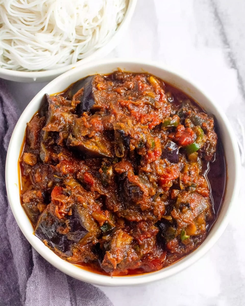 A white bowl filled with a rich, chunky dark reddish-brown stew made of thick slices of eggplant covered in a thick, textured sauce with visible small bits of green peppers and tomatoes. The sauce looks slightly oily and glossy, showing layers of spices and cooked ingredients well-mixed. The bowl rests on a white cloth with a white marbled surface underneath, and part of another white bowl with thin white noodles is visible in the top left corner. Photo taken with an iphone --ar 4:5 --v 7