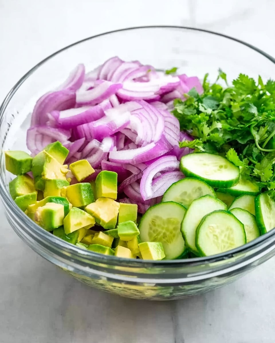 The image shows a clear glass bowl on a white marbled surface, filled with layers of fresh chopped vegetables. On the left side, there are thin slices of purple-red onion. Next to the onions, toward the bottom left, are small green cubes of avocado. To the right, bright green cucumber slices with light seeds are placed. A small bunch of green cilantro leaves sits behind the cucumber slices. The bowl is arranged with the ingredients separated in groups, showing a mix of colors and textures. Photo taken with an iphone --ar 4:5 --v 7
