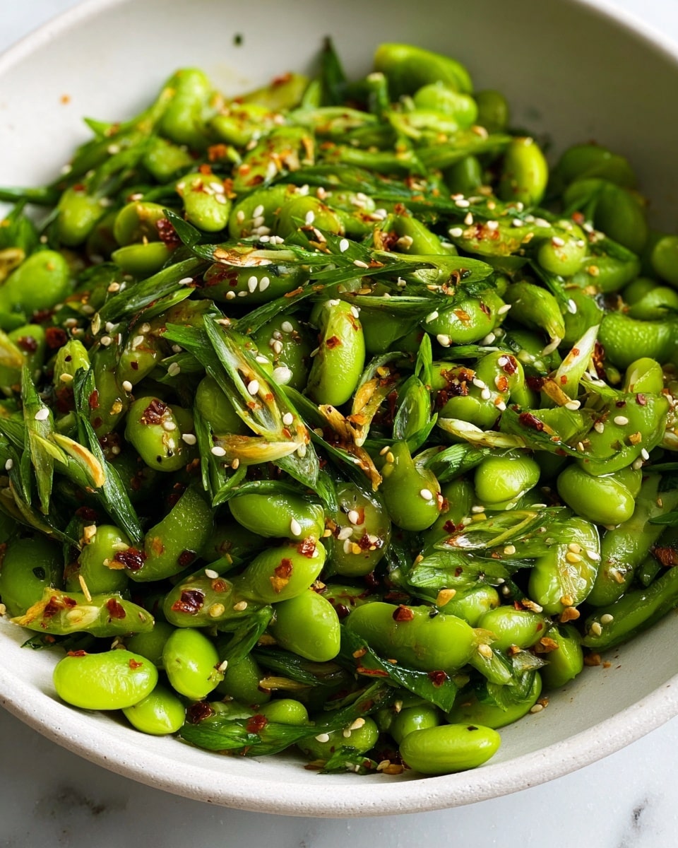 A close-up view of a vibrant green salad in a white bowl on a white marbled surface, showing two main layers: the base layer is bright green edamame beans scattered thickly, mixed with thinly sliced green onions adding a fresh texture, and the top layer is sprinkled with small white sesame seeds and reddish chili flakes providing a contrast in color and a hint of spice, with a light sheen of dressing that makes the ingredients glisten, slightly oily but fresh and lively, the salad looks mixed but with clear separate layers visible; photo taken with an iphone --ar 4:5 --v 7