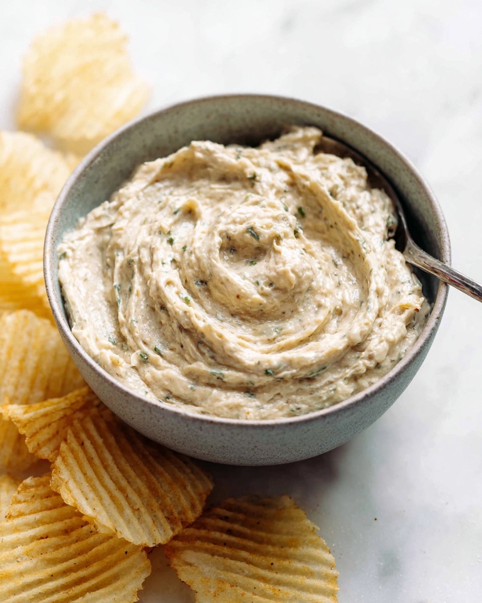 A close-up of a dark bowl filled with creamy, thick dip mixed with shredded pieces that are light brown and green herbs, showing a smooth and textured surface. A woman's hand is holding a large, ridged potato chip dipped halfway in the creamy mixture, positioned at the right edge of the image. The background is a white marbled texture, giving a clean and bright look. photo taken with an iphone --ar 4:5 --v 7