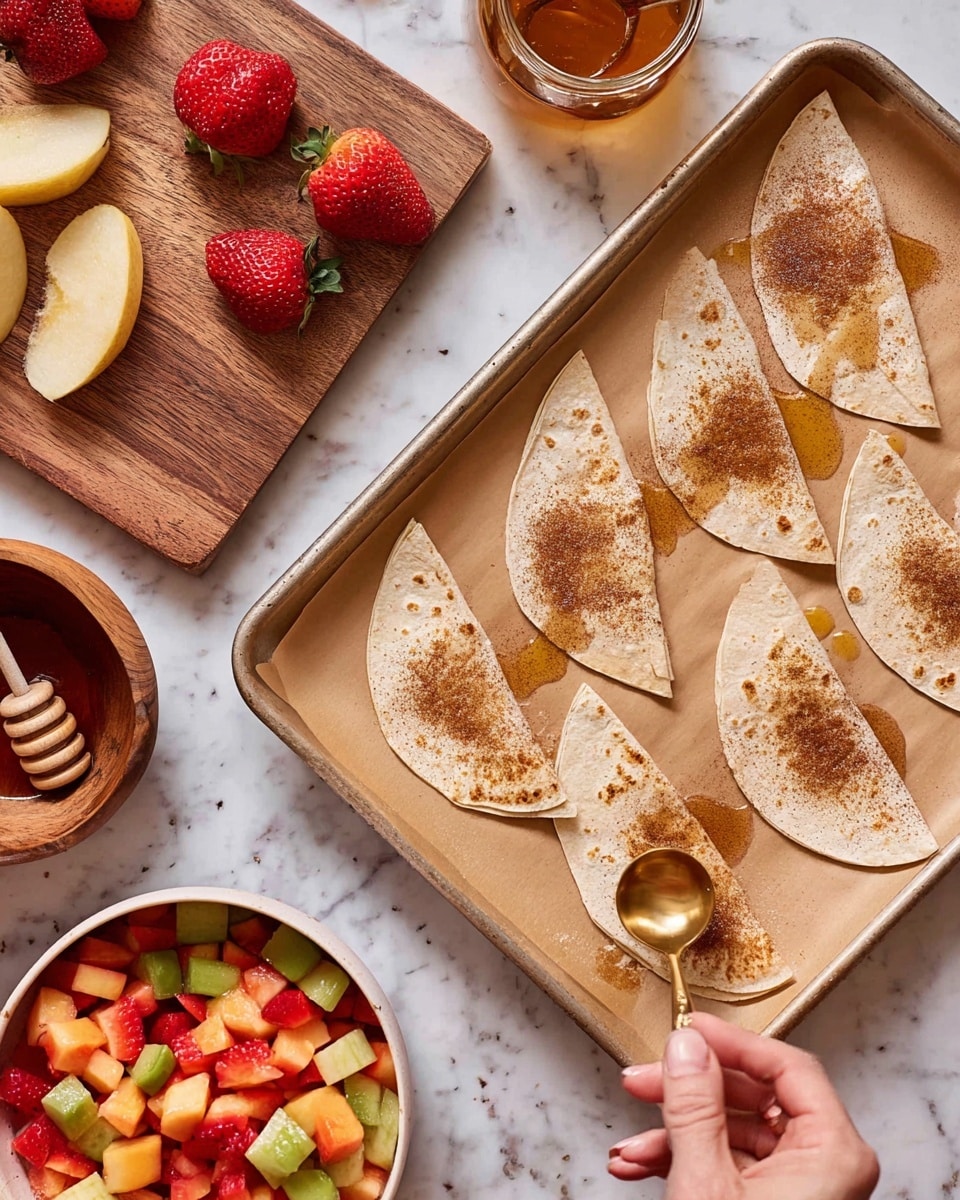 The image shows a baking tray lined with brown parchment paper, with eight tortilla pieces cut into quarter-circle shapes spread out on it. Each tortilla piece has a light drizzle of oil and is sprinkled with a brown powder, likely cinnamon or similar spice. A woman's hand is holding a small golden spoon, adding the powder to one of the tortillas. To the left of the tray are a wooden cutting board with strawberries and a slice of cantaloupe, a small wooden bowl filled with the same brown powder, and a glass jar of honey with a honey dipper inside. Below the tray is a large white bowl filled with diced mixed fruit, featuring red, green, and orange pieces, and a spoon inside it. The background is a white marbled surface. photo taken with an iphone --ar 4:5 --v 7