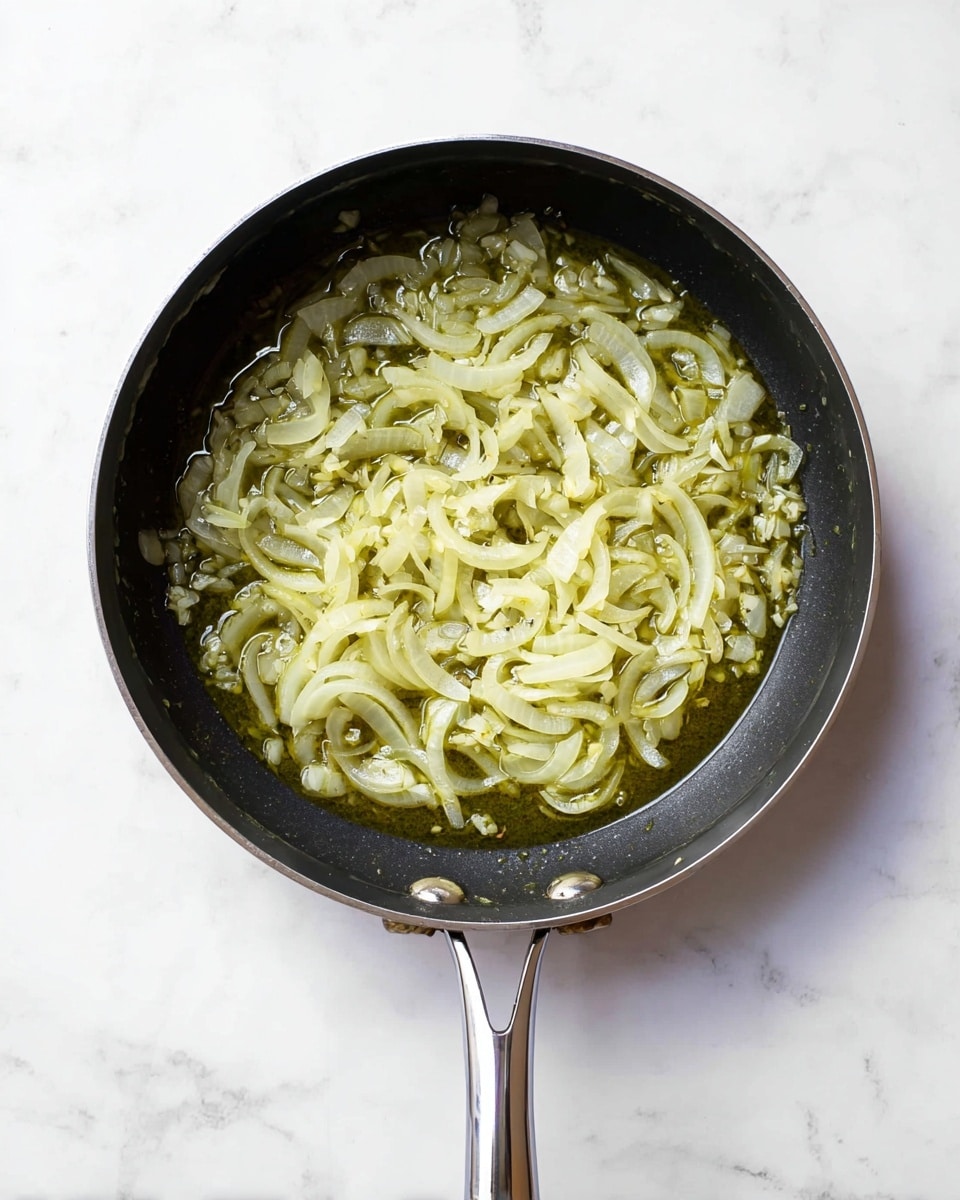 A top view of a black metal pan with a silver handle, filled with thinly sliced translucent onions and minced garlic cooking in olive oil. The onions are soft and wet, floating in the shiny greenish-golden oil that covers the bottom of the pan. The background is a white marbled surface. Photo taken with an iphone --ar 4:5 --v 7