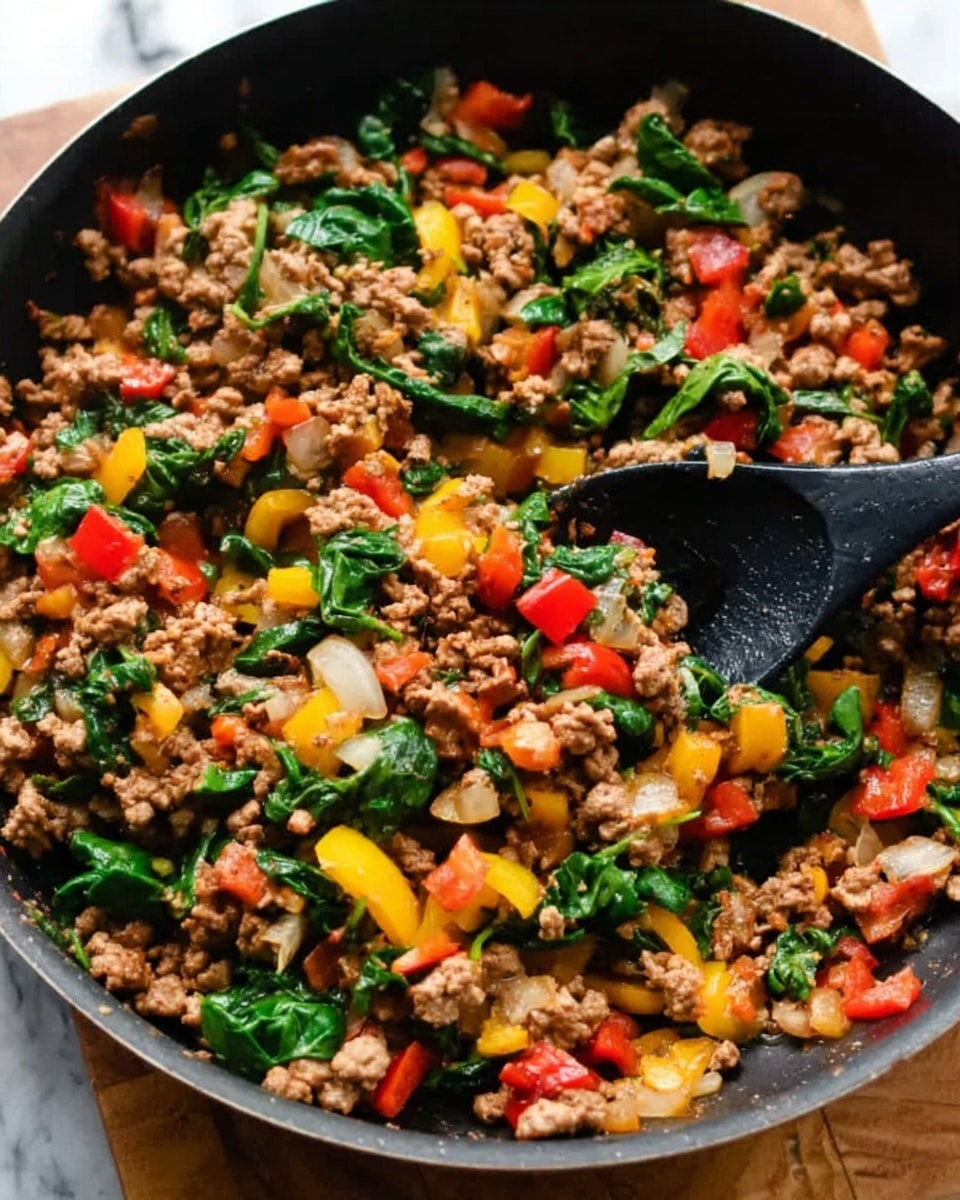 The image shows a close-up of a black pan filled with cooked ground meat mixed with chopped vegetables. The dish has many layers including brown cooked meat, bright red and yellow bell pepper pieces, fresh green spinach leaves, and some white onion slices. A black spoon is scooping some of the mixture from the pan. The pan is placed on a wooden surface with a white marbled texture visible around it. The colors are rich and natural, showing a warm, hearty dish photo taken with an iphone --ar 4:5 --v 7