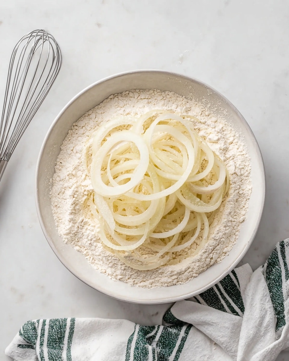 A white bowl filled with a thick layer of off-white flour sits on a white marbled surface. On top of the flour, there are several layers of raw onion rings that are pale yellow and translucent, loosely placed over one another in a circle. To the left of the bowl is a metal whisk, and to the lower right corner, there is a white cloth with dark green stripes. The lighting is bright and natural, highlighting the soft texture of the flour and the thin, smooth surface of the onion rings. photo taken with an iphone --ar 4:5 --v 7