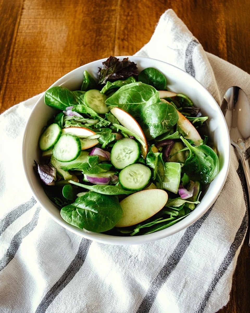A white bowl filled with fresh green salad layers including leafy spinach and lettuce at the base, sliced light green cucumbers scattered on top, thin slices of red apple adding a hint of red, and small pieces of purple onion adding contrast. The salad rests on a white cloth with thin black stripes, all placed on a wooden surface. Photo taken with an iphone --ar 4:5 --v 7