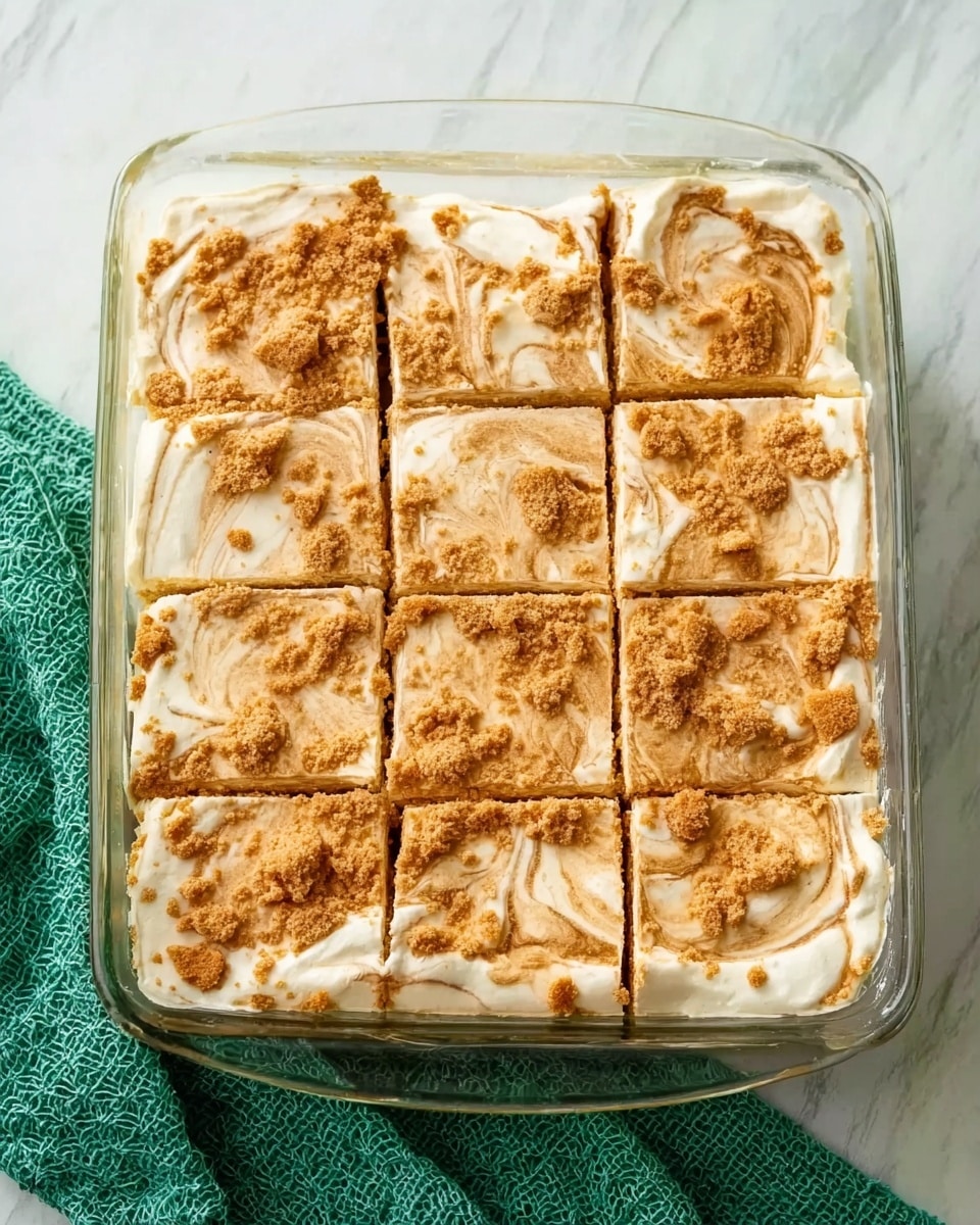 A rectangular clear glass baking dish filled with a creamy dessert that is cut into nine squares. The top layer is light beige with swirled patterns of a darker brown, giving a marbled effect. The texture looks smooth and soft with some crumbly bits scattered on top. The dish is placed on a white marbled surface with a green textured cloth partially visible on the side. Photo taken with an iphone --ar 4:5 --v 7