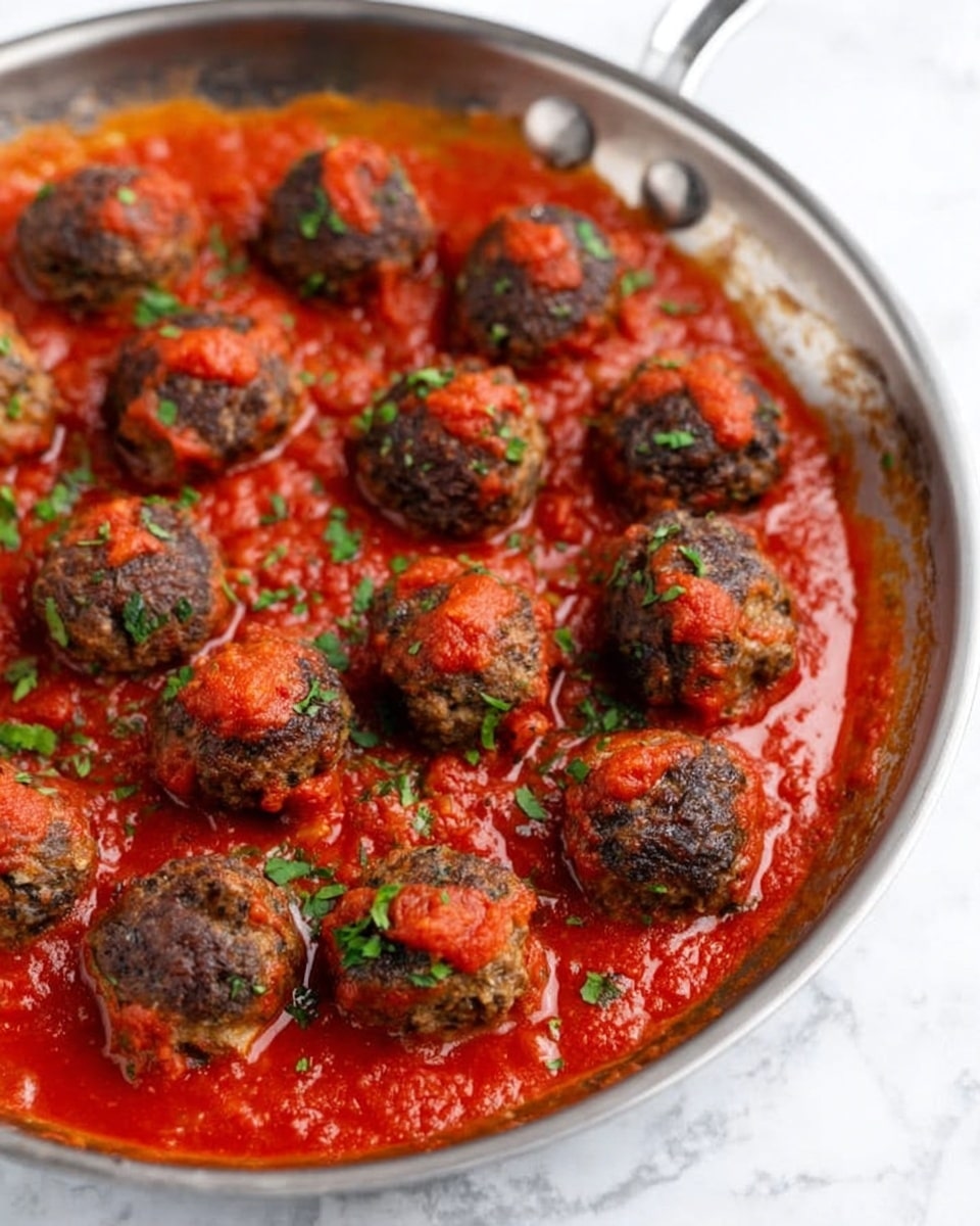 The image shows a silver metal pan filled with small, round meatballs covered in red tomato sauce. The meatballs are dark brown with a slightly rough texture, and each one is topped with a small dollop of bright red sauce. The sauce covers the bottom of the pan and surrounds the meatballs, with some small green herbs sprinkled across the dish. The pan sits on a white marbled surface. photo taken with an iphone --ar 4:5 --v 7