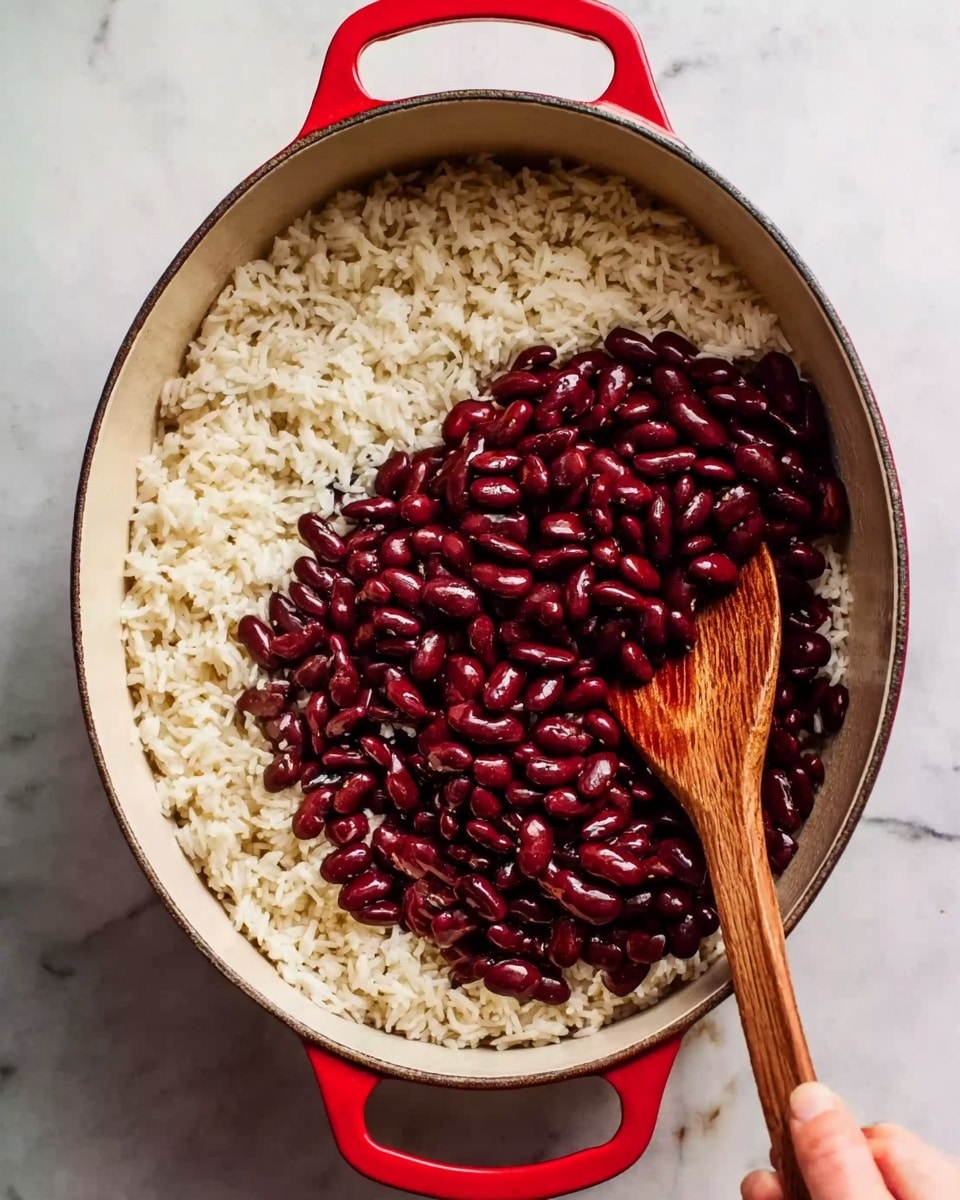 The image shows a white oval pot with red handles placed on a white marbled surface. Inside the pot, there is a layer of cooked white rice evenly spread out as the base layer. On top of the rice, there is a large pile of dark red kidney beans forming the middle layer in the center. A wooden spoon is resting on the right side inside the pot, partially touching both the rice and beans, with a woman’s hand holding the pot’s handle from the left side. The photo taken with an iphone --ar 4:5 --v 7