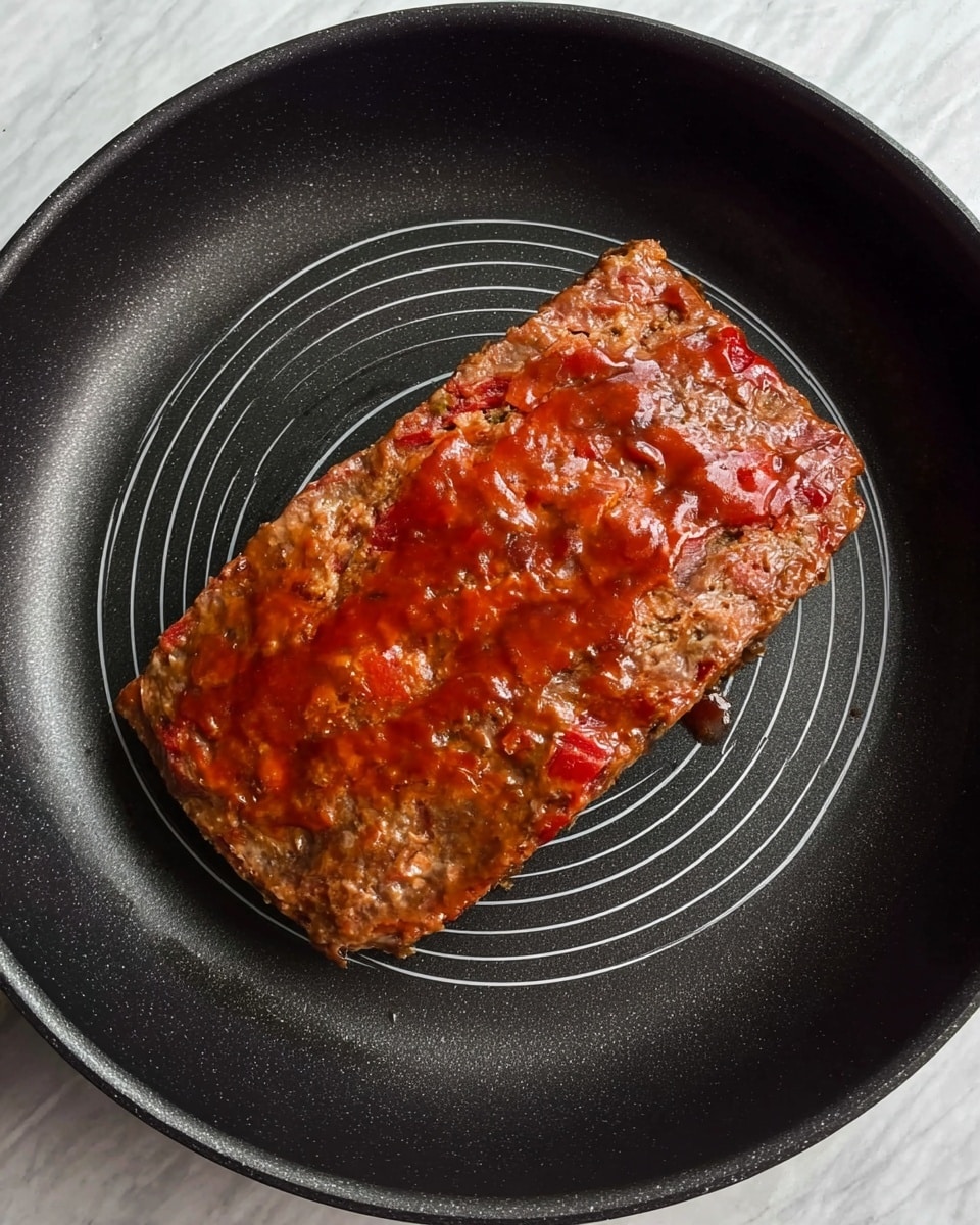A rectangular, brown meatloaf sits in the center of a black frying pan with evenly spaced gray lines forming a circular pattern underneath. The top layer of the meatloaf is covered with a slightly shiny, reddish sauce spread unevenly, creating a textured surface with some streaks and small patches of different red shades. The pan rests on a white marbled surface. photo taken with an iphone --ar 4:5 --v 7