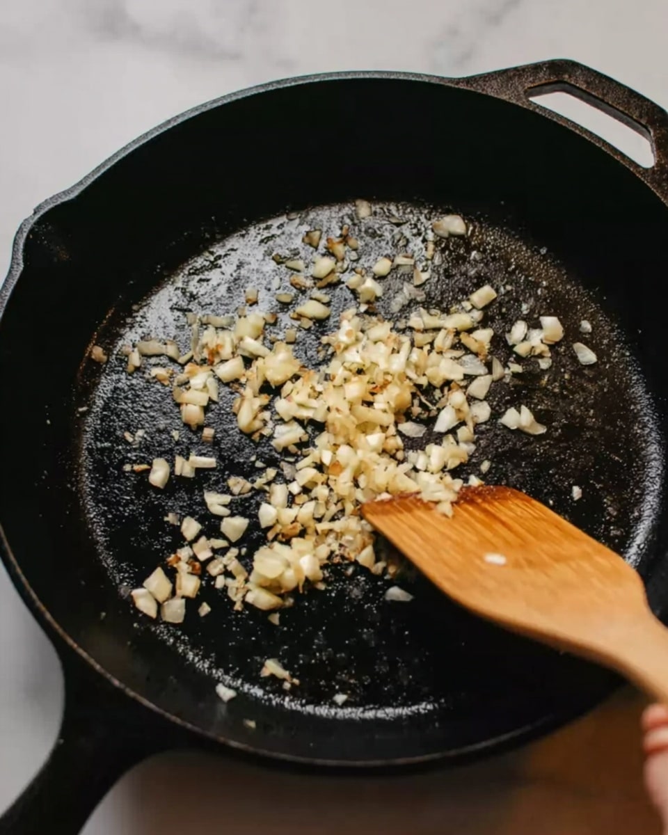 A black cast iron pan is shown from above, with small pieces of chopped garlic evenly spread across its surface. A wooden spatula, held by a woman's hand, is stirring the garlic, which is light golden in color with a slightly translucent texture. The background is a white marbled surface. Photo taken with an iphone --ar 4:5 --v 7