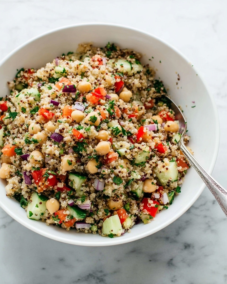 A white bowl filled with a mix of quinoa, chickpeas, diced red bell peppers, chopped cucumbers, finely chopped red onions, and small green parsley leaves all mixed together. The quinoa colors are light beige with some darker grains, giving a textured look. Chunky pieces of red bell pepper and cucumber are spread evenly throughout, with pale round chickpeas adding bigger, smooth dots. The red onions provide small purple bits, and the green parsley adds fresh green specks all over. A silver spoon is partially inside the bowl on the right side. The bowl rests on a white marbled surface. Photo taken with an iphone --ar 4:5 --v 7