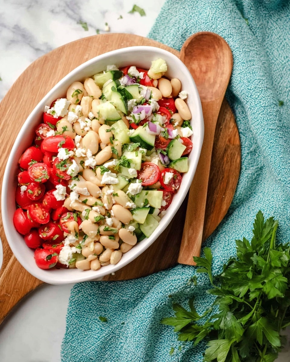 The image shows a white oval bowl filled with a colorful bean salad on a wooden round board, which is placed on a turquoise textured cloth over a white marbled surface. The salad has three main layers: the first layer has creamy white beans, the second layer has small green cucumber cubes and bright red cherry tomato halves, and the third layer is made up of scattered white cheese bits and chopped green herbs. A wooden spoon rests next to the bowl on the board, and fresh green parsley lies near the bottom right corner of the image. Photo taken with an iphone --ar 4:5 --v 7