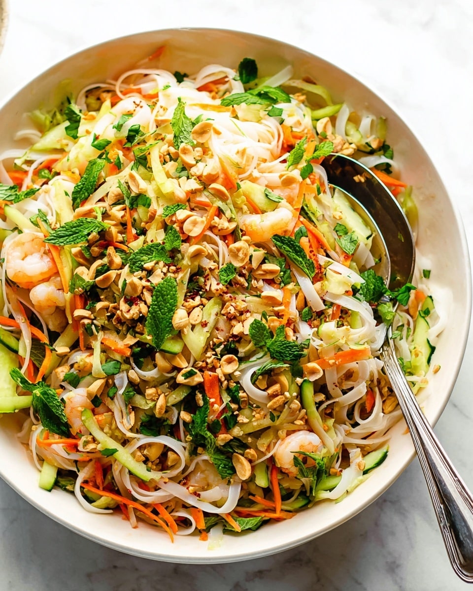 A large white bowl sits on a white marbled surface filled with a colorful noodle salad. The bottom layer has flat white rice noodles, soft and slightly glossy. Mixed through are small pieces of pale shrimp and crunchy green cucumber slices. Thin orange carrot strips and small white cabbage chunks add texture, scattered with fresh green herbs like cilantro and mint leaves. Light brown crushed peanuts and black pepper flecks are sprinkled on top, and a shiny metal spoon rests inside the bowl, ready to serve. photo taken with an iphone --ar 4:5 --v 7