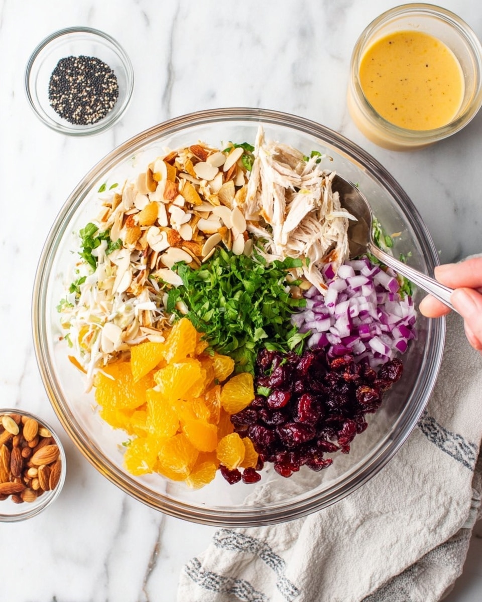 A clear glass bowl on a white marbled surface contains seven distinct layers arranged in separate sections. Starting from the top moving clockwise, there are light brown crispy strips, white shredded chicken, bright orange mandarin pieces, light beige sliced almonds, finely chopped purple onions, dark red dried cranberries, and a central pile of fresh green herbs. A small clear glass bowl with black seeds sits nearby on the left, while a glass of light orange dressing is placed to the right. A woman's hand is holding a silver serving spoon inside the bowl, and a white and gray cloth is partially visible underneath. photo taken with an iphone --ar 4:5 --v 7