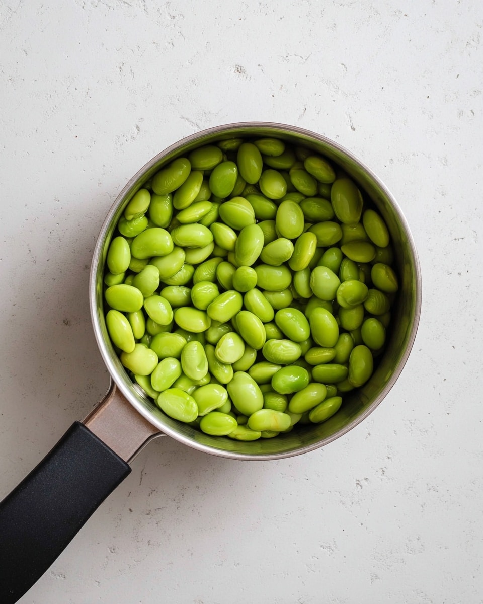 A small silver pot filled with a single layer of bright green edamame beans, each bean smooth and shiny, tightly packed inside the pot. The pot is on a white marbled surface with tiny gray speckles, and the pot's black handle extends out to the left side. There is a clear view from the top, showing only the green beans inside the pot photo taken with an iphone --ar 4:5 --v 7
