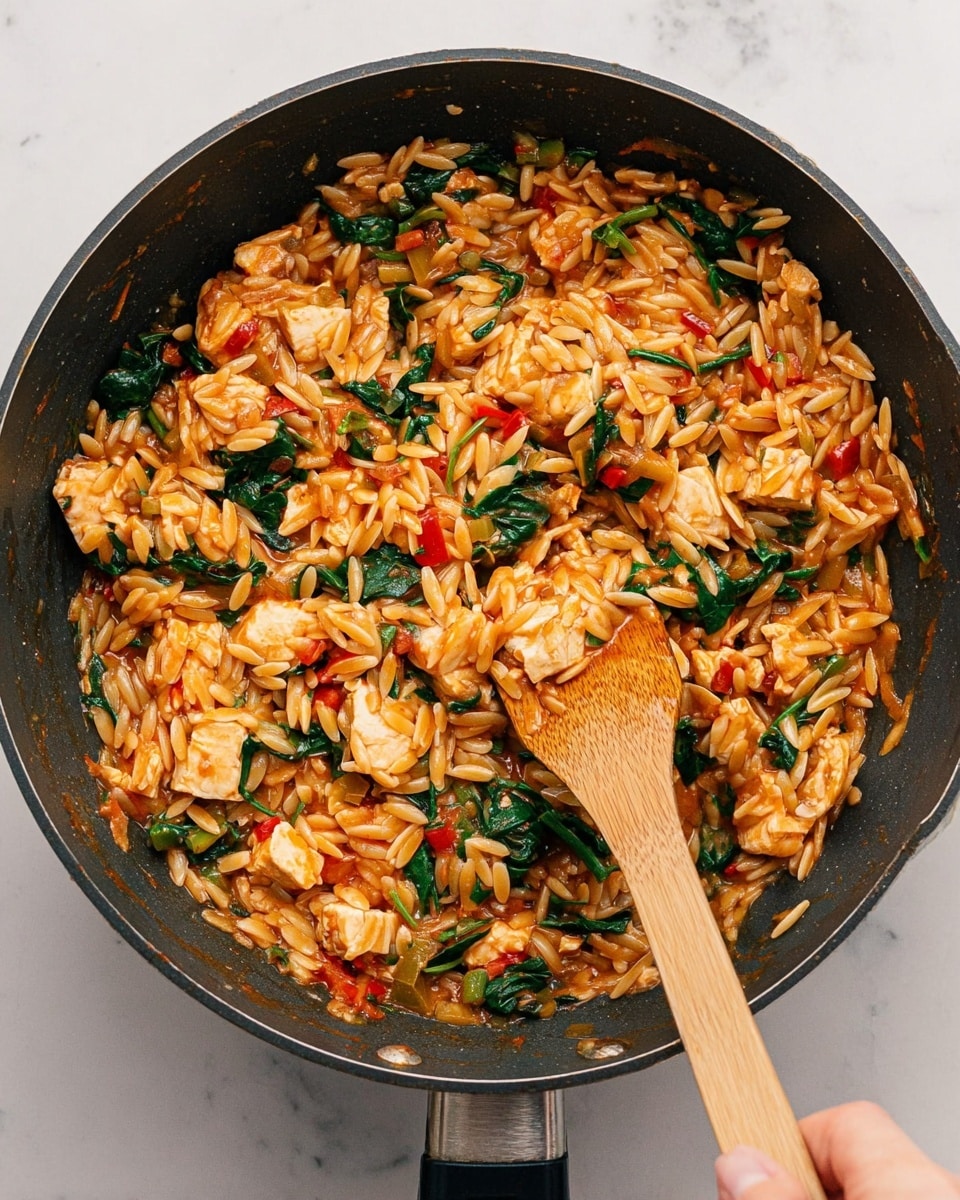 The image shows a dark cooking pan filled with a mixed dish of orzo pasta, diced white meat pieces, cooked spinach leaves, and small pieces of green and red bell peppers all coated in a reddish-orange sauce. The orzo pasta is oval and light brown, mingling with the chunky white meat and vibrant green spinach scattered throughout. A wooden spatula is stirring the mixture inside the pan, held by a woman's hand at the bottom right corner. The background is a white marbled surface. photo taken with an iphone --ar 4:5 --v 7