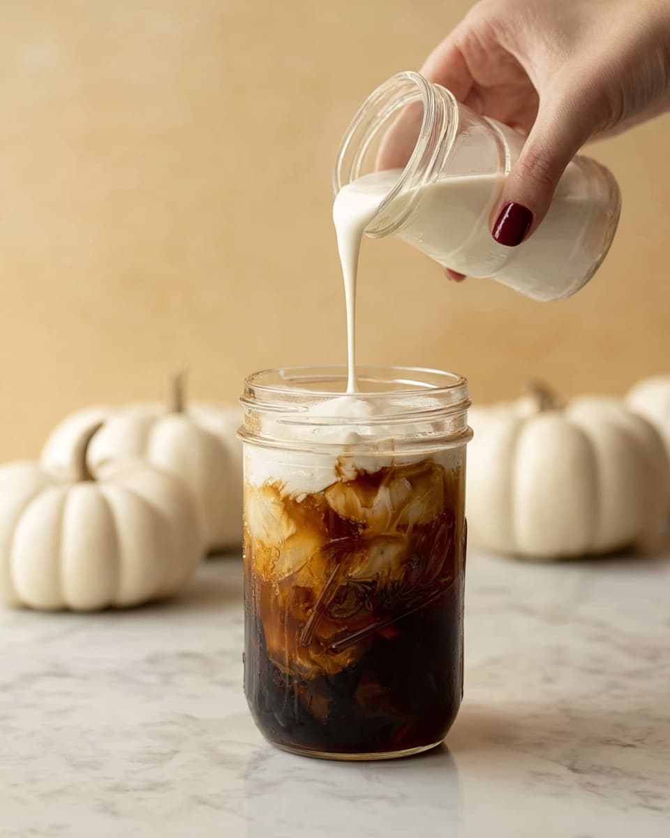 A woman's hand with dark red nail polish is pouring thick white cream from a small clear jar into a transparent glass mason jar filled with dark brown iced coffee and visible ice cubes. The cream flows in a smooth layer on top of the coffee, creating a foamy white cap over the dark liquid. The mason jar sits on a white marbled surface. In the blurred background, there are three small white pumpkins, adding a light cream color contrast against the warm beige wall behind. Photo taken with an iphone --ar 4:5 --v 7