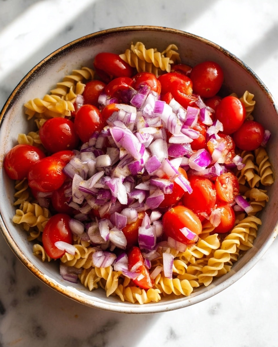 The dish shows a white round bowl filled with three layers: the bottom layer has yellow spiral pasta, the middle layer contains bright red halved cherry tomatoes, and the top layer is made up of small diced purple and white onion pieces scattered unevenly over the tomatoes. The bowl sits on a white marbled surface with a soft light coming from the upper right, highlighting the fresh colors of the ingredients photo taken with an iphone --ar 4:5 --v 7
