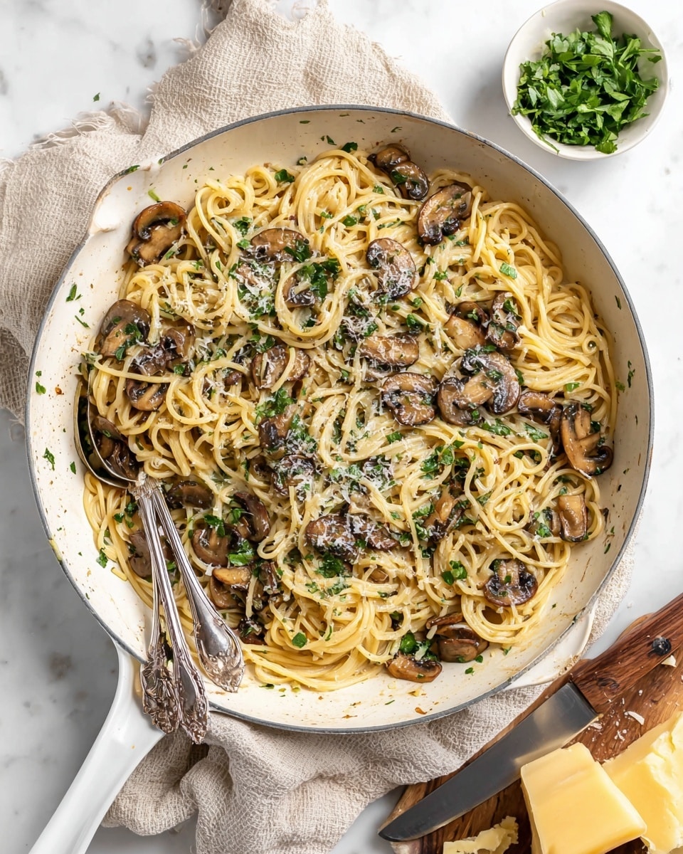 A white skillet filled with creamy spaghetti pasta, mixed with golden brown sautéed mushroom slices scattered evenly throughout. The pasta has a smooth, glossy texture and is topped with chopped green herbs and some grated cheese, adding a touch of white and green on top. Two vintage silver utensils rest inside the skillet, placed slightly on the left side. The skillet sits on a beige cloth over a white marbled surface, with a small white bowl of fresh green herbs in the upper right corner and a wooden-handled knife next to chunks of light yellow cheese near the bottom right. The overall scene has a bright and clean look, highlighting the colors and textures of the dish. photo taken with an iphone --ar 4:5 --v 7