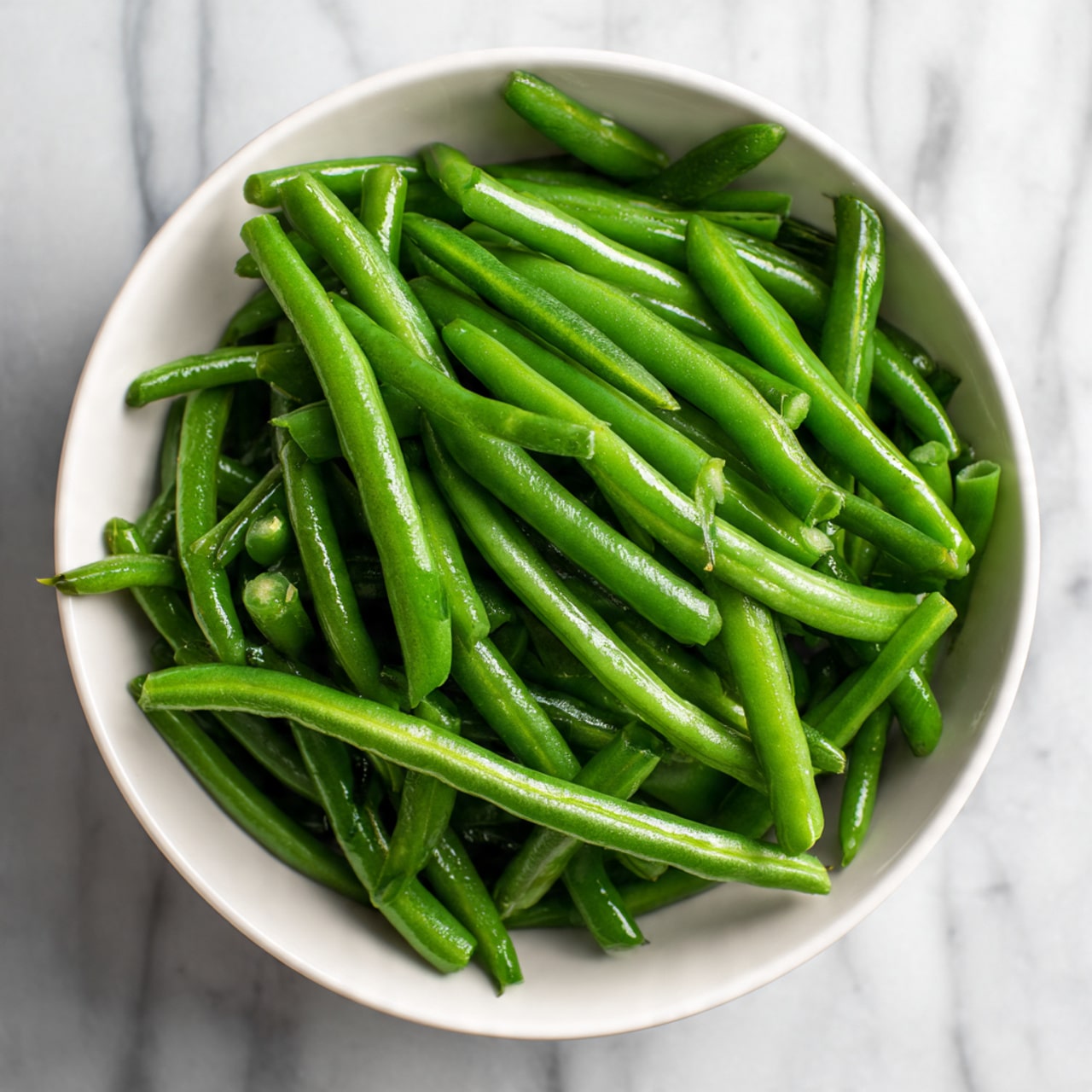 A white bowl filled with fresh green beans that are sliced lengthwise, showing the smooth outer skin and the inside with small beans visible. The green beans are piled casually inside the bowl with some overlapping. The bowl is placed on a surface with a white marbled texture. The lighting is natural, highlighting the fresh green color and smooth texture of the green beans photo taken with an iphone --ar 4:5 --v 7