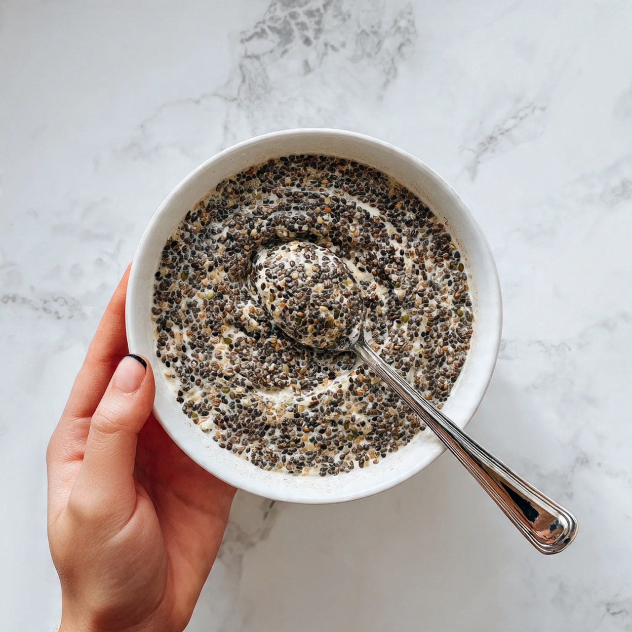 A large white bowl is filled with a thick mixture of seeds showing layers of small black, white, and green seeds with a rough texture, swirled in a circular pattern. A spoon with a metallic shine is partially dipped into the mixture towards the right side, held by a woman's hand. Another woman's hand is at the bottom left holding the bowl against a white marbled surface background. The bowl has a smooth inside with some dark speckles on the rim. Photo taken with an iphone --ar 4:5 --v 7