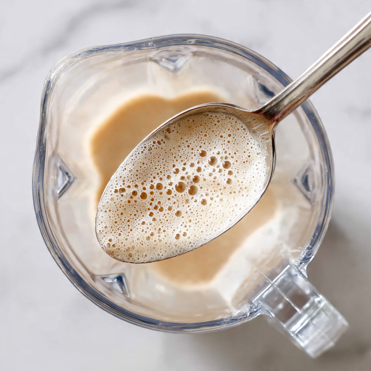 A close-up image shows a silver spoon lifting a light beige, bubbly liquid mixture inside a clear glass blender jar. The mixture has small frothy bubbles on its smooth surface, and the jar has visible corners and edges. The background is a white marbled texture, softly blurred to highlight the jar and spoon in the center. Photo taken with an iphone --ar 4:5 --v 7