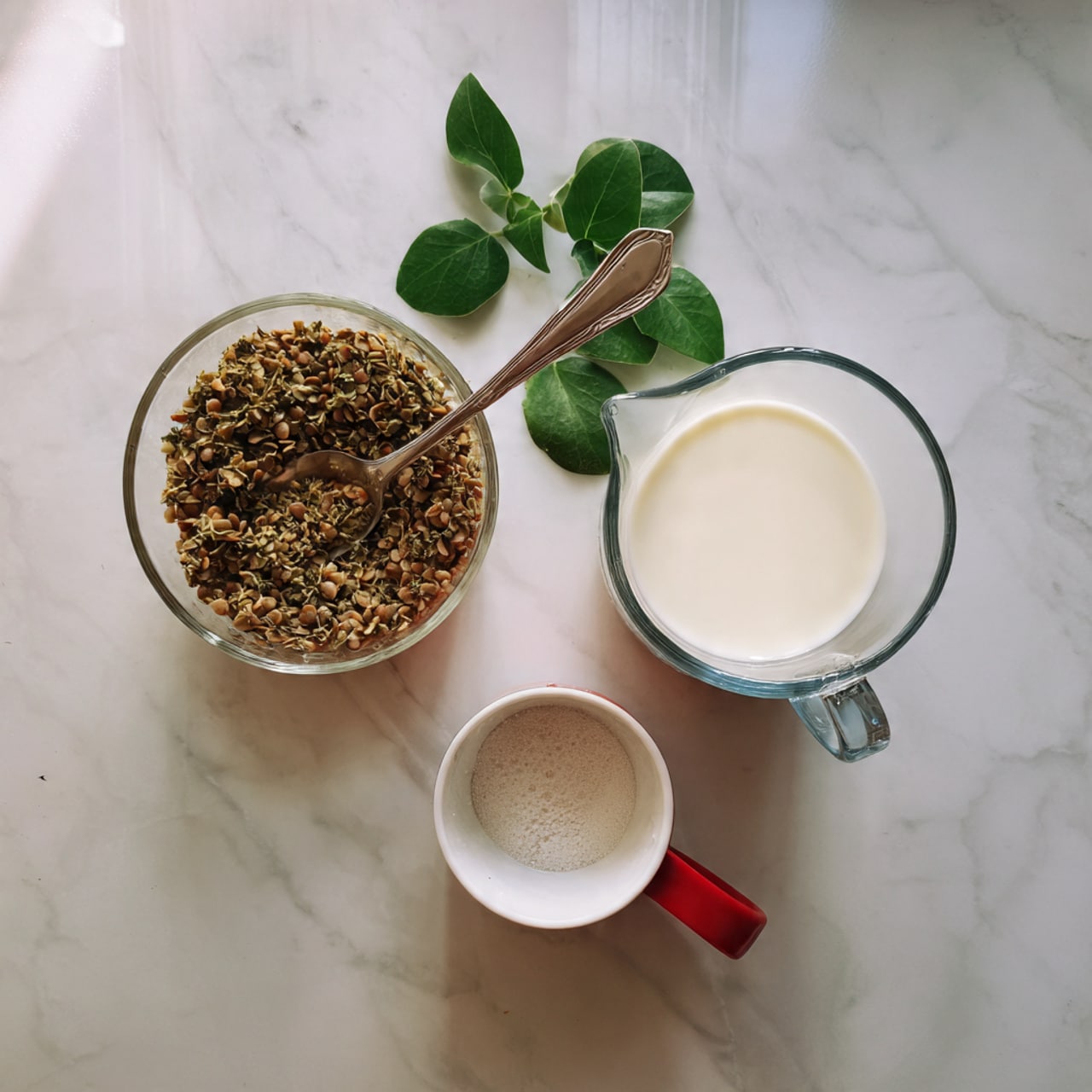 The image shows three main items on a white marbled surface. On the left, there is a clear glass bowl with a thick greenish mixture that looks like cooked lentils, with a silver spoon inside the bowl. Above this bowl, there are a few fresh green leaves placed as decoration. To the right, there is a clear measuring cup filled with white milk, its smooth texture clearly visible. Above the milk is a small white cup with a red handle filled with a white powdery ingredient. The setup is clean and simple, with the focus on the glass containers and their contents, all placed neatly on the light surface. photo taken with an iphone --ar 4:5 --v 7