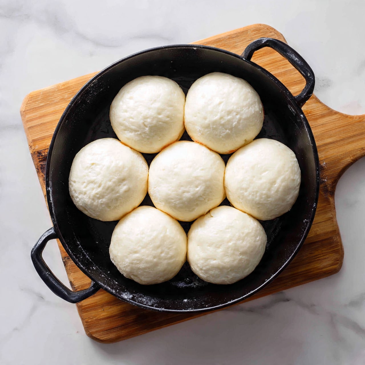 A black cast iron pan holding eight small white dough rounds that are soft and smooth, spread evenly across the pan. The pan sits on a white marbled textured surface with a wooden cutting board partially visible underneath. The dough pieces are simple, pale, and thick, with a slight puffiness showing before baking. photo taken with an iphone --ar 4:5 --v 7