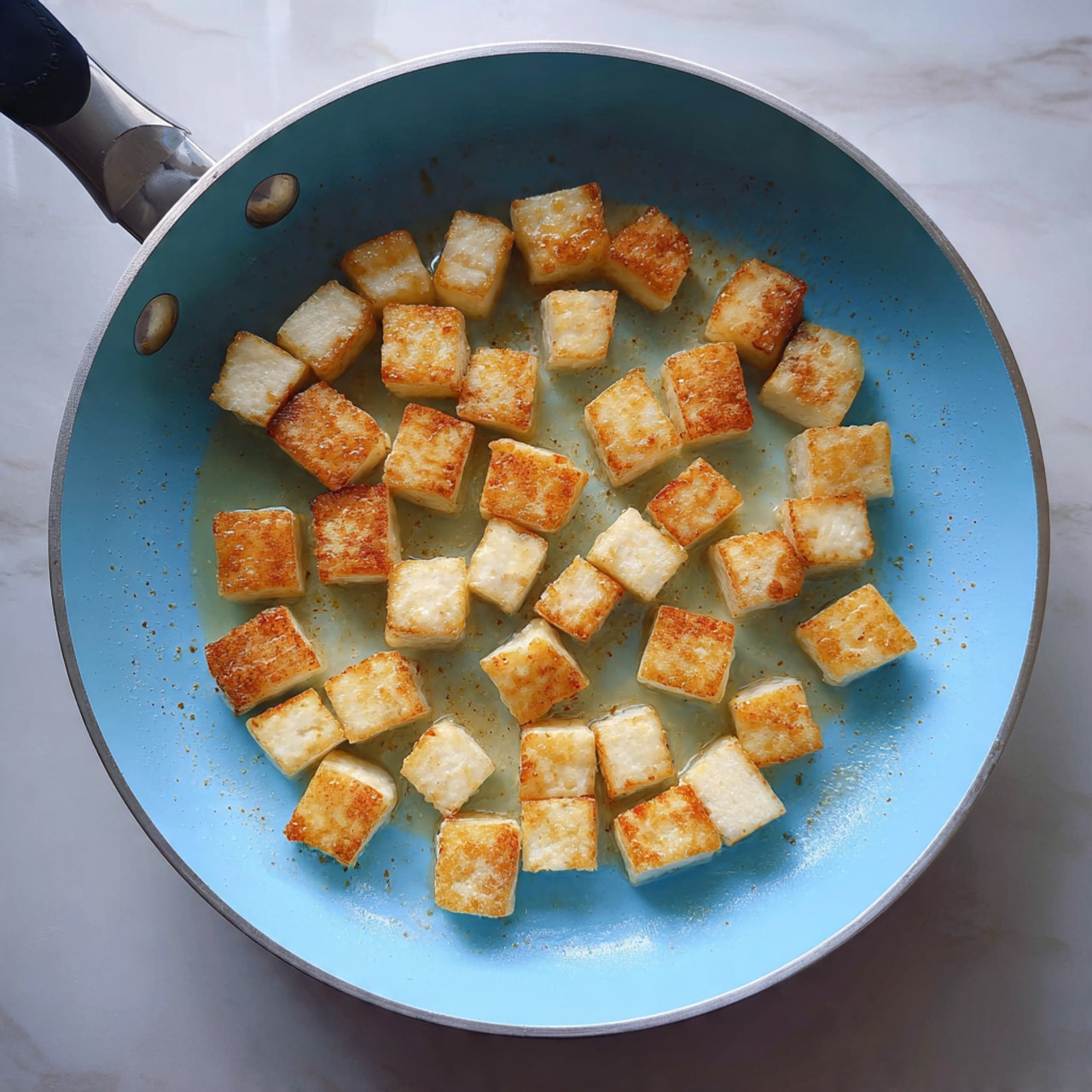 The image shows a light blue frying pan filled with evenly spaced, small cubes of tofu being cooked. Each tofu cube has a light golden brown color on the top surface, showing a slightly crispy texture while the sides remain pale and soft. The pan holds a small amount of oil, which gives a shiny, moist look to the tofu pieces. The background is a white marbled surface. Photo taken with an iphone --ar 4:5 --v 7