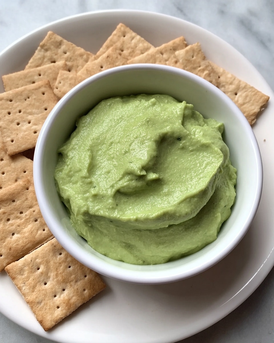 A small white bowl filled with a smooth, thick green dip that has a slightly creamy texture and some soft ridges on its surface, surrounded on a white plate by light brown square crackers with small perforations and a slightly grainy texture, all placed on a white marbled surface. photo taken with an iphone --ar 4:5 --v 7