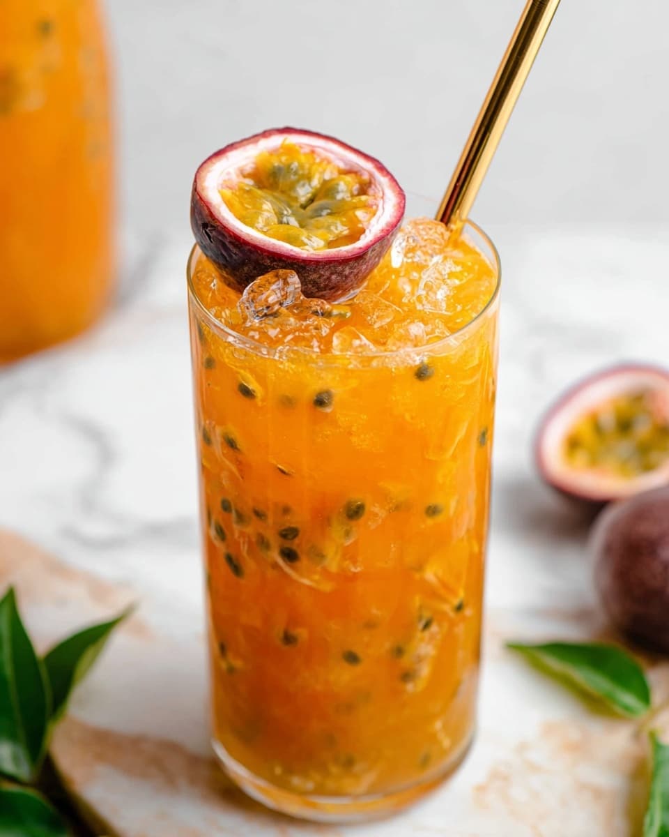 A tall clear glass filled with a bright orange drink that has visible black seeds and crushed ice floating on top, creating a textured and icy layer. On top of the drink, there is a halved passion fruit with yellow pulp and black seeds inside the purple shell, sitting slightly on the side. A golden metal straw is placed inside the glass, leaning on the left edge. The glass is set on a white marbled surface with a blurred out orange drink and some green leaves in the background, along with a whole passion fruit beside the glass. Photo taken with an iphone --ar 4:5 --v 7