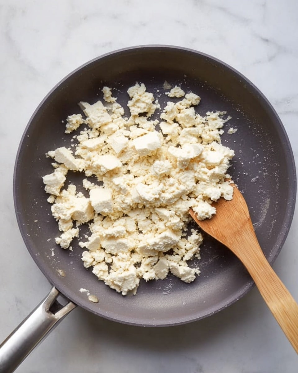 The image shows a dark gray frying pan placed on a white marbled surface, filled with crumbled white tofu pieces evenly spread inside. A wooden spatula with a smooth texture is resting in the pan, partly lifting some of the tofu, showing the mixture's fluffiness. The tofu has a soft, crumbly texture with some small bits scattered around the larger chunks. The handle of the pan is silver, extending towards the bottom left of the picture. The lighting is bright and natural, highlighting the white color of the tofu and the soft shadows inside the pan. photo taken with an iphone --ar 4:5 --v 7