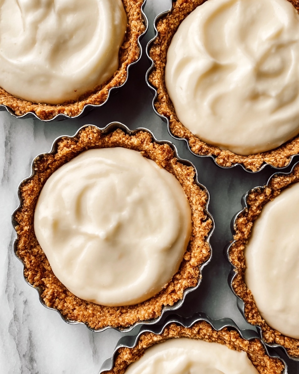 The image shows four small tarts in shiny metal tart pans, arranged close together on a white marbled surface. Each tart has two layers: a rough, crumbly, golden-brown crust on the outer edge and bottom, and a smooth, creamy, pale beige filling that looks soft and slightly textured on top. The filling has gentle swirls, showing it was spread with a soft motion, and it fills the crust just to the rim. The metal pans have scalloped edges that nicely frame the crust. Photo taken with an iphone --ar 4:5 --v 7