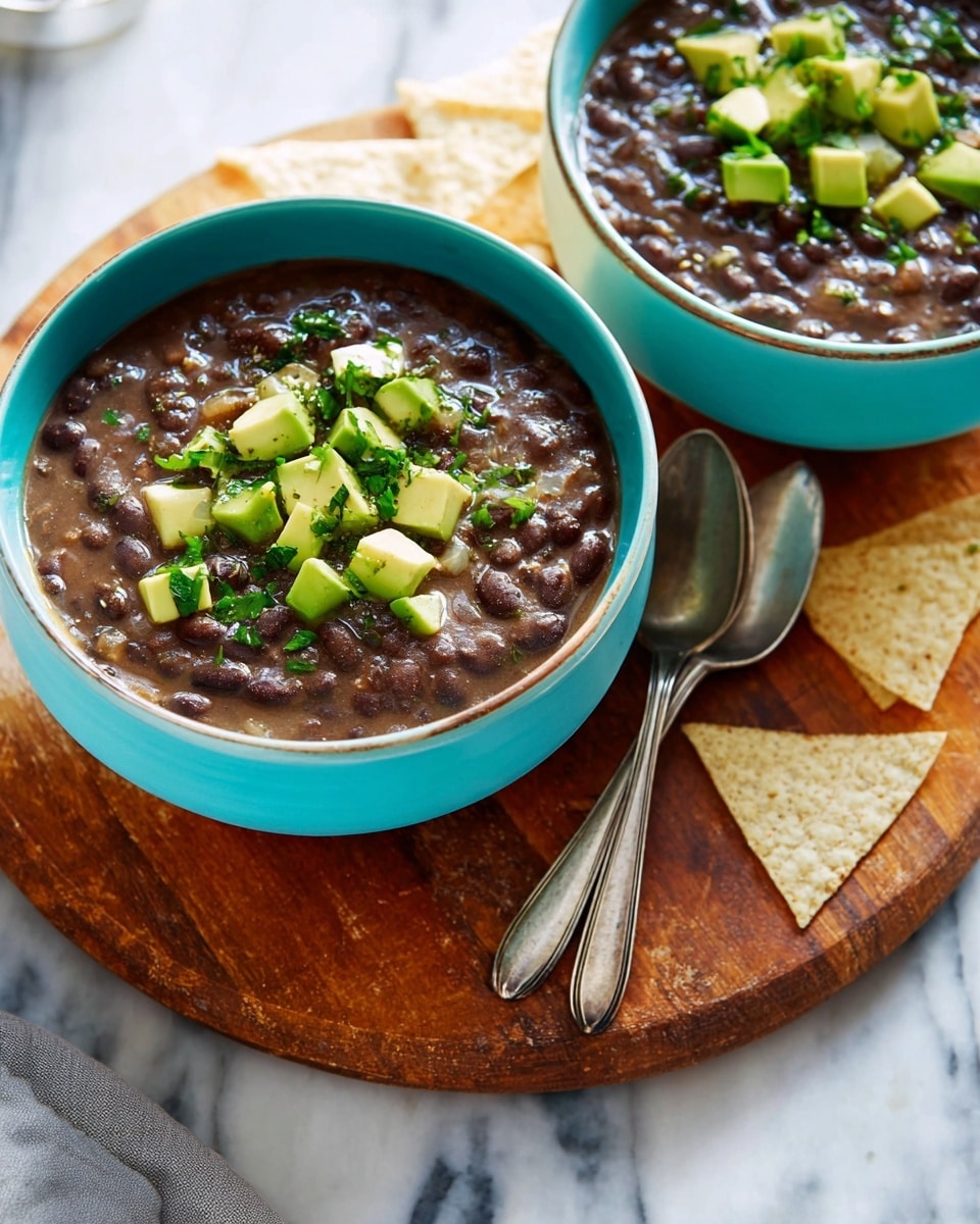 Two turquoise bowls filled with thick, dark brown beans topped with light green avocado cubes and small pieces of chopped green herbs. The bowls are placed on a round wooden board, with two silver spoons resting on the board between them. Parts of white, triangular tortilla chips are visible around the board on a white marbled surface. The scene is well lit, showing textures of creamy beans and fresh avocado, photo taken with an iphone --ar 4:5 --v 7