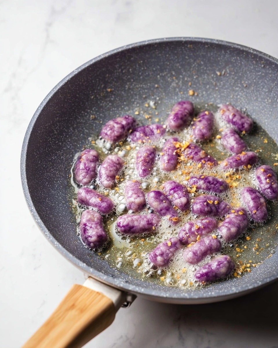 A grey speckled frying pan with a light wooden handle contains small purple gnocchi pieces frying in bubbling butter. The gnocchi appear soft and slightly wrinkled, scattered evenly across the pan's surface with some golden bits of seasoning sprinkled on them. The butter has small foam bubbles, showing it is hot and sizzling. The background is a white marbled texture. photo taken with an iphone --ar 4:5 --v 7