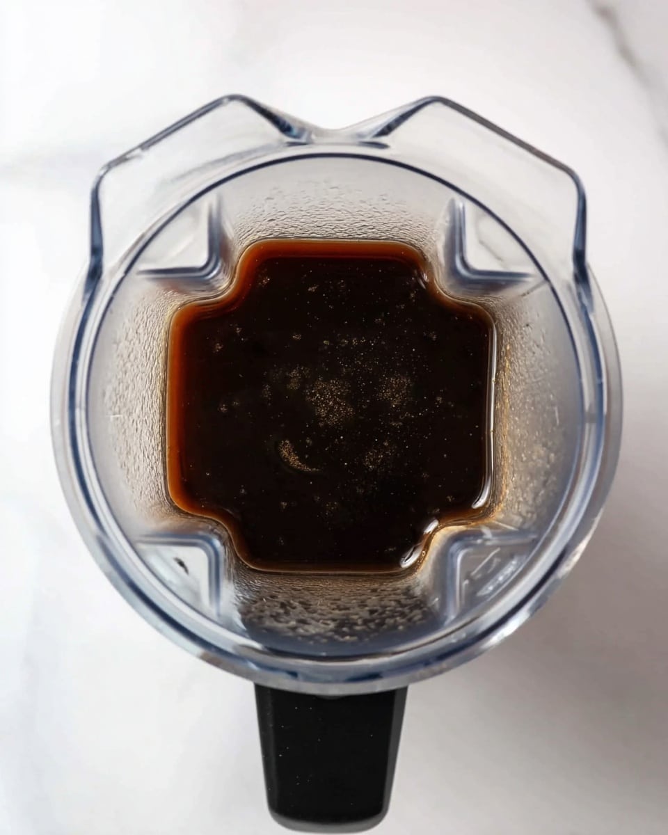 The image shows a top view inside a clear blender with sharp edges and ridges, filled with a small amount of dark brown liquid at the bottom. The blender is placed on a white marbled surface, and the black handle of the blender is visible on the left side. The liquid inside looks smooth and shiny, with some small bubbles or spots on the surface. Photo taken with an iphone --ar 4:5 --v 7