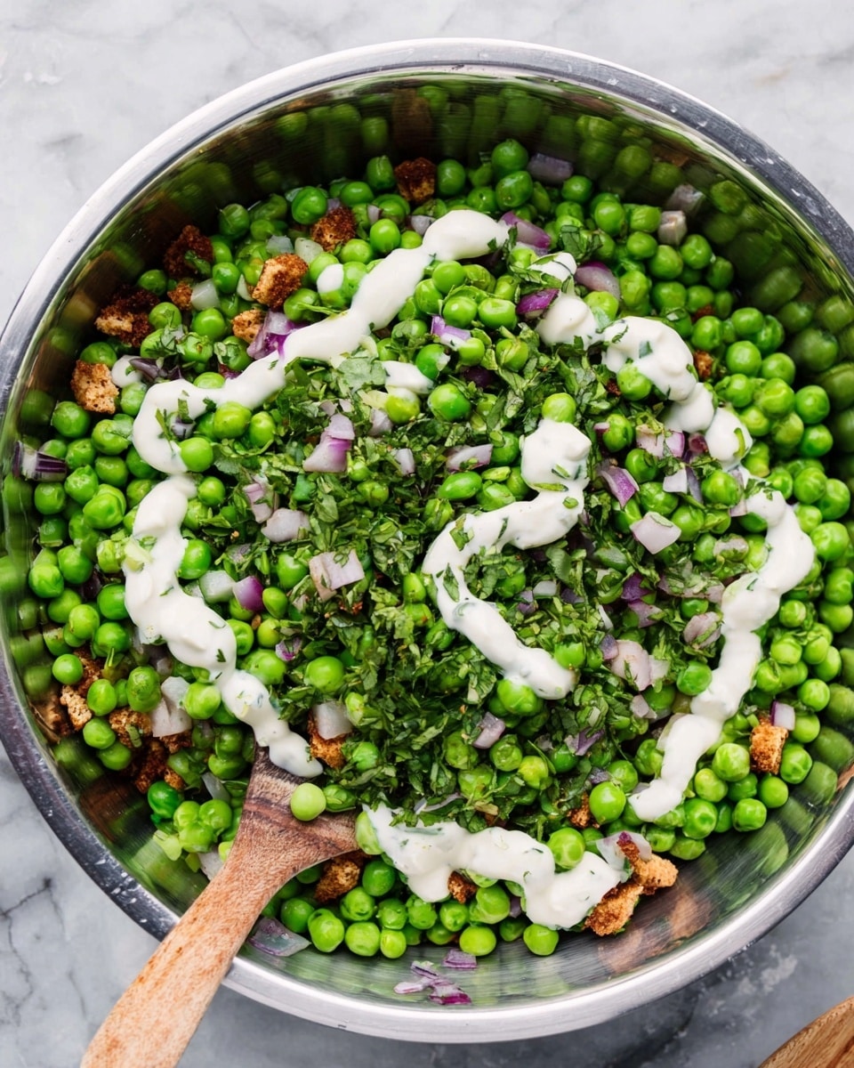 A stainless steel bowl filled with bright green peas as the main layer, mixed with small pieces of light brown crumbled tofu and chopped fresh green herbs scattered throughout. There are also small purple onion pieces spread across the peas. A white creamy sauce is drizzled on top in a zigzag pattern, adding contrast against the green peas. The bowl is on a white marbled surface, and a wooden spoon is resting inside the bowl, partially visible. photo taken with an iphone --ar 4:5 --v 7