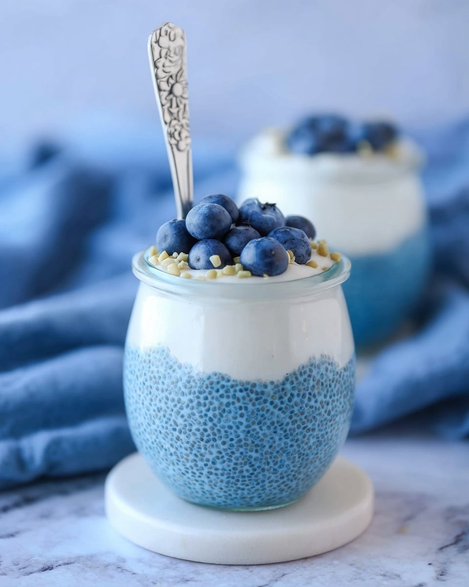 The image shows a clear glass jar filled with two layers of chia pudding. The bottom layer is bright blue with visible chia seeds giving it a textured, bumpy look. Above it is a smooth, white creamy layer. The top surface is decorated with fresh, round blueberries and small light-colored seeds scattered around. A silver spoon with a floral pattern on the handle stands upright in the jar. The jar sits on a small, white coaster against a soft blue fabric background on a white marbled surface. Another similar jar is blurred in the background. Photo taken with an iphone --ar 4:5 --v 7