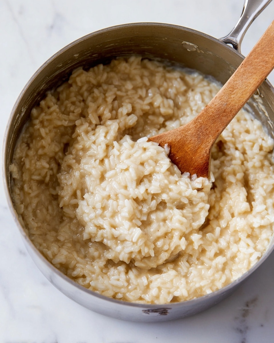 The image shows a close-up of a stainless steel pot filled with creamy risotto. The risotto is pale beige in color with a soft, thick texture, and the grains of rice are visible, swollen and coated in sauce. A wooden spoon is partly submerged in the risotto, scooping some of the rice, and its handle extends out from the pot towards the bottom right of the frame. The pot is placed on a white marbled surface that adds a clean, bright contrast to the warm tone of the risotto. The scene is simple and focused on the creamy rice dish photo taken with an iphone --ar 4:5 --v 7
