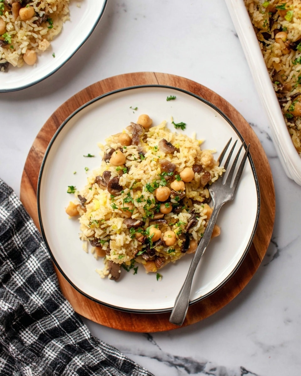 A white round plate with a thin black rim holding a layered serving of rice mixed with chickpeas, small chopped mushrooms, and bits of cooked onion, all topped with small pieces of fresh green herbs. The rice is light yellow with scattered textures of beige chickpeas and dark brown mushrooms, sitting on a wooden circular board against a white marbled surface. A silver fork rests on the plate’s right side, and a black and white checked cloth lies beside the board. Part of a white baking dish with more of the rice mixture is visible in the background. Photo taken with an iphone --ar 4:5 --v 7