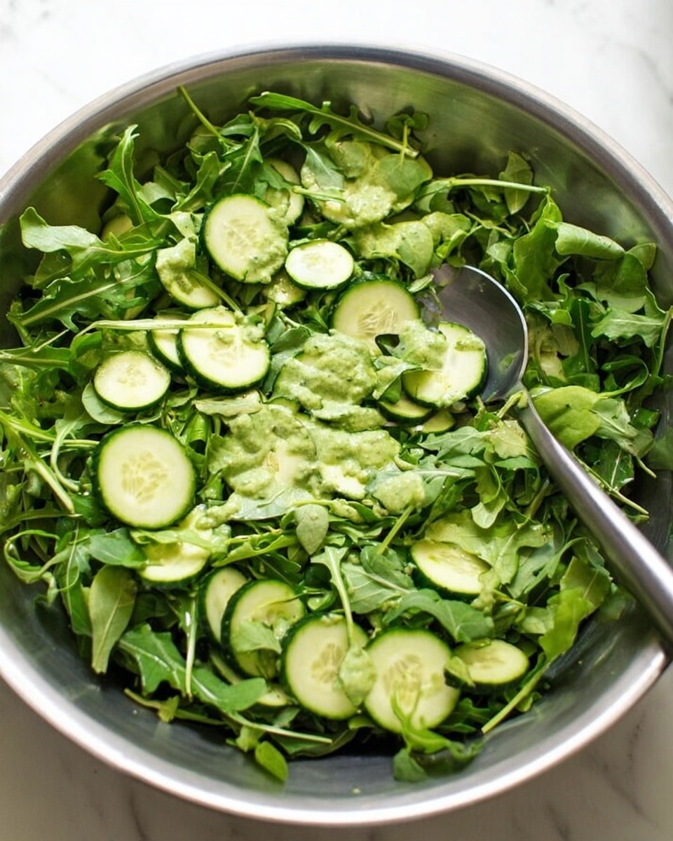 A large shiny metal bowl filled with fresh green arugula leaves forming the base layer, topped with thinly sliced round cucumber pieces scattered evenly over the greens. A light green creamy dressing is drizzled unevenly on top, adding texture and a hint of gloss. A silver spoon rests inside the bowl on the right side, ready for serving, and everything sits on a white marbled surface. photo taken with an iphone --ar 4:5 --v 7