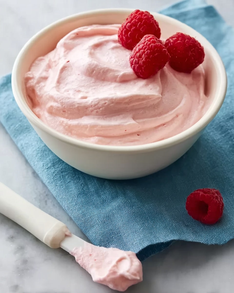 A white bowl filled with smooth, light pink cream that has a soft, whipped texture, topped with three fresh red raspberries on one side. Next to the bowl lies a white spatula with some of the same pink cream on its head, resting on a blue cloth. One fresh raspberry is placed on the white marbled surface near the spatula. The overall setup has soft lighting, highlighting the creamy texture and fresh fruit. photo taken with an iphone --ar 4:5 --v 7