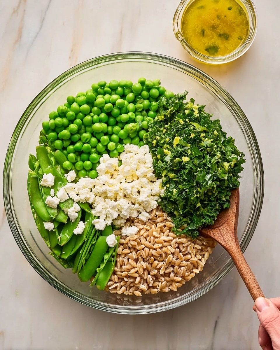 A clear glass bowl is filled with four distinct layers of ingredients arranged in separate sections: bright green snap peas and peas on the top left, vibrant green chopped kale on the top right, white crumbled cheese below the kale, and light brown cooked farro below the peas. The bowl is set on a white marbled surface. Next to the bowl on the upper right is a small jar filled with a yellow dressing and a spoon inside it. A woman's hand is gently holding a wooden spoon inside the bowl. photo taken with an iphone --ar 4:5 --v 7