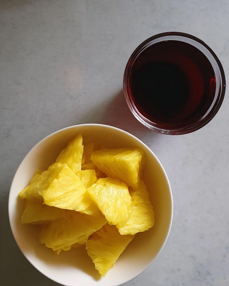 The image shows a white bowl filled with bright yellow pineapple pieces, arranged loosely inside. Above the bowl, there is a clear glass cup filled with a dark reddish-brown liquid. The background is a smooth white marbled texture. photo taken with an iphone --ar 4:5 --v 7