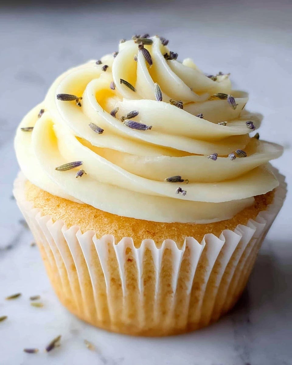 A single cupcake is shown on a white marbled surface, with a golden-brown base in a white cupcake liner. The top layer is made of creamy pale yellow frosting, swirled in a smooth and thick rosette shape. Small dried lavender buds are sprinkled gently on the frosting, adding subtle texture and dark greenish-gray color spots. The frosting looks soft and rich, forming clear petal-like layers. photo taken with an iphone --ar 4:5 --v 7