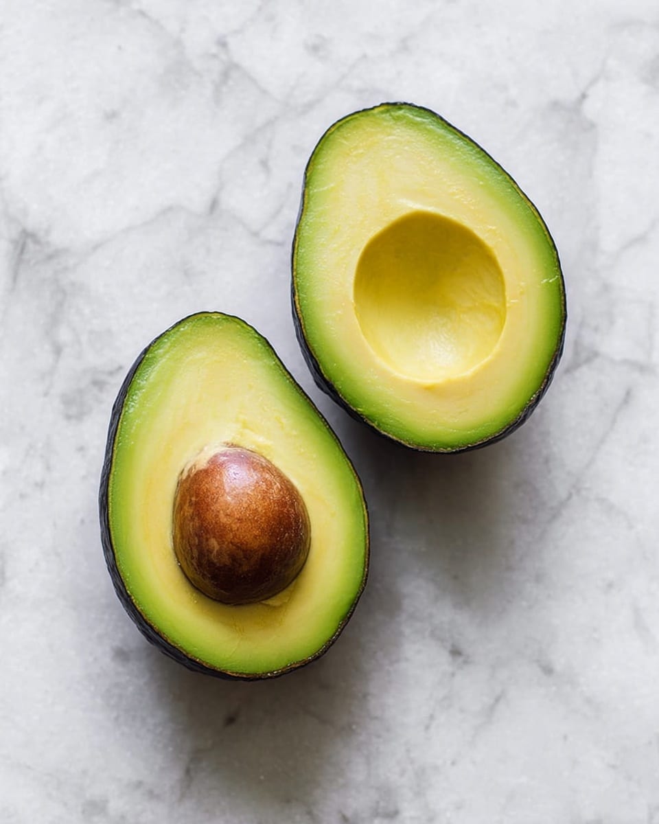 Two halves of a ripe avocado are placed on a white marbled surface. The left half shows a smooth, yellow-green inside where the seed has been removed, with a darker green edge near the skin. The right half has a large, round, brown seed sitting perfectly inside the bright green flesh that fades into yellow toward the center, surrounded by a dark green peel. photo taken with an iphone --ar 4:5 --v 7